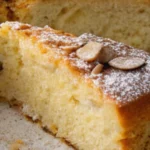 A slice of moist almond nut cake topped with sliced almonds and powdered sugar.