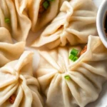 Steamed pork chive dumplings with pleated wrappers served on a white plate with soy dipping sauce.