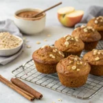 Applesauce muffins with oat topping cooling on a wire rack beside cinnamon sticks and apple sauce