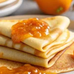 Close-up of Crêpes Suzette with golden crêpes folded neatly, topped with rich orange sauce on a ceramic plate.