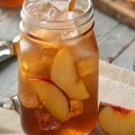 A mason jar filled with sweet tea peaches, packed with fresh peach slices and ice cubes, sitting on a rustic wooden table.