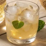 A glass of coconut infused rum served over ice with a fresh mint or leaf garnish, beside a bottle of rum and a coconut on a wooden table.