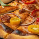 Close-up of grilled pizza slices with charred crust, yellow cherry tomatoes, red onions, sausage, and fresh herbs.
