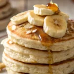 Stack of fluffy oatmeal pancakes topped with fresh banana slices and drizzled with maple syrup.